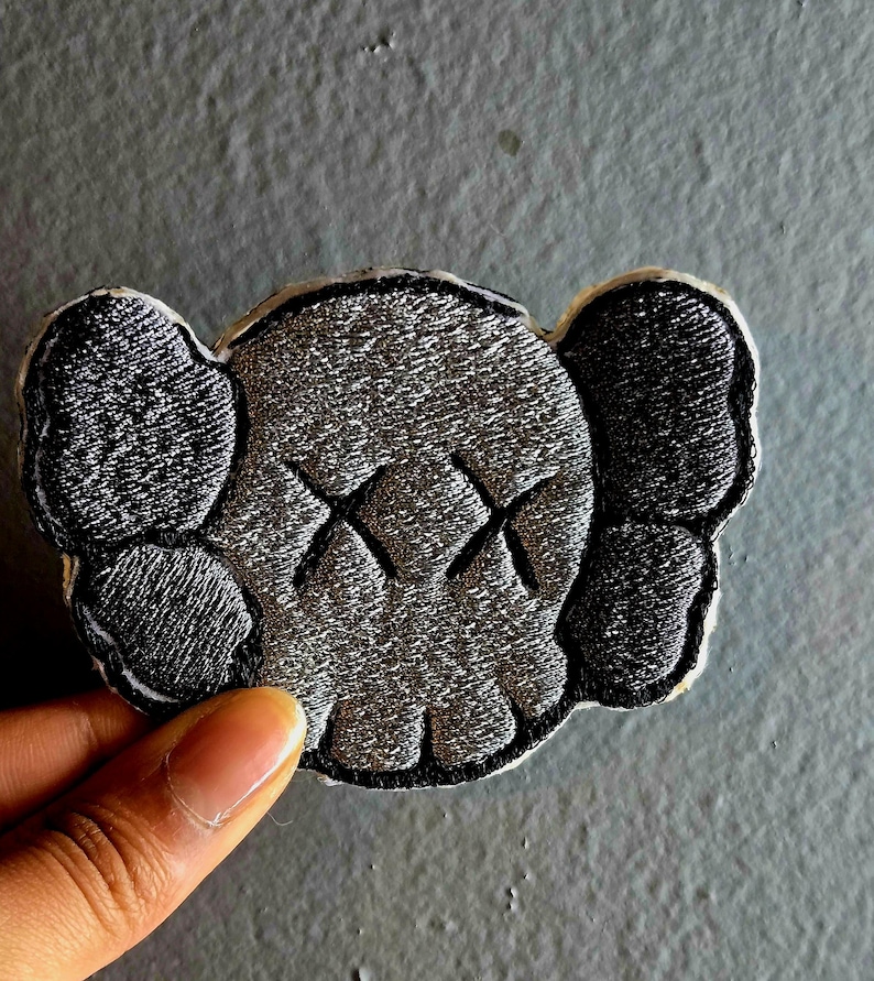 May include: Embroidered patch featuring a stylized skull design with crossed-out eyes. The patch has a gray and black color scheme with a white border. The design includes large ear-like shapes on either side of the skull.