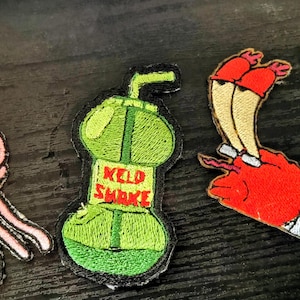 May include: Three embroidered patches: a pink jellyfish, a green Kelp Shake, and a red and tan character. The Kelp Shake patch has the text "Kelp Shake".