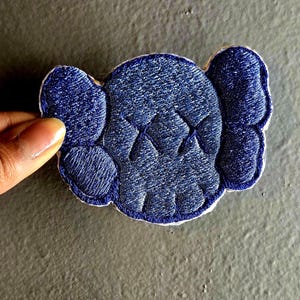 May include: A navy blue embroidered patch in the shape of a cartoon skull with X-shaped eyes. The patch has a white border and is held by a hand against a gray background. The design features a textured, stitched appearance.