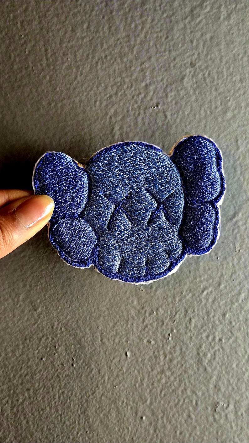 May include: A navy blue embroidered patch in the shape of a cartoon skull with X-shaped eyes. The patch has a white border and is held by a hand against a gray background. The design features a textured, stitched appearance.