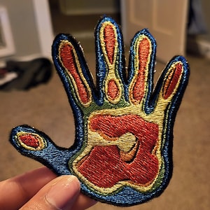 May include: Embroidered patch in the shape of a hand, featuring a vibrant design. The palm is red, surrounded by yellow, green, and blue accents. The patch has a black border and is likely for clothing or accessories.