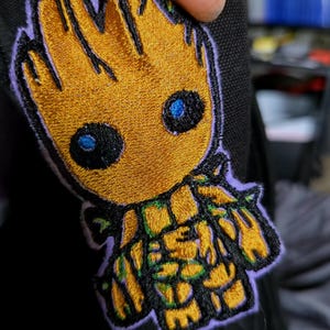 May include: Embroidered patch featuring a stylized character with a golden-yellow body, black eyes, and blue pupils. The patch has a purple border and is likely for application on clothing or accessories.