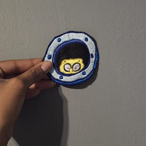 May include: Embroidered patch featuring a cartoon character peeking through a blue and silver porthole. The character has yellow skin and large, wide eyes. The patch is circular and has a detailed, stitched design.