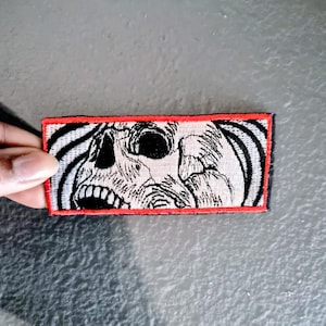 May include: Embroidered patch featuring a detailed, black and white skull design with an open mouth and hollow eye sockets. The skull is set against a background of swirling black and white lines, framed by a red border.