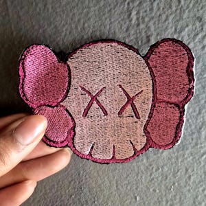 May include: Embroidered patch featuring a stylized skull design with crossed-out eyes and large ear-like shapes. The patch is primarily light pink with darker pink accents and a black border. The design is reminiscent of pop art.