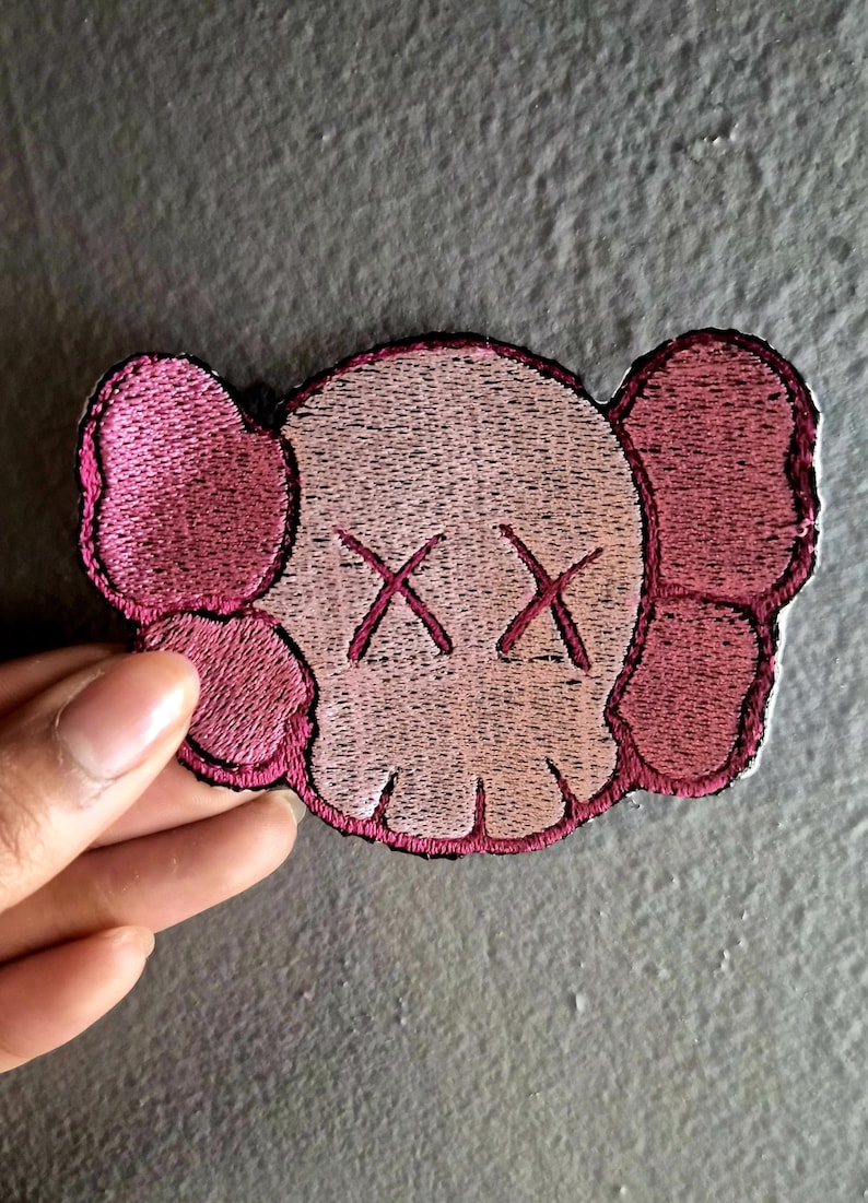 May include: Embroidered patch featuring a stylized skull design with crossed-out eyes and large ear-like shapes. The patch is primarily light pink with darker pink accents and a black border. The design is reminiscent of pop art.