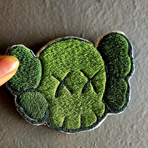 May include: Embroidered green patch featuring a stylized skull with X-shaped eyes and rounded ears. The patch has a dark green border and is set against a neutral background. The design is detailed with textured stitching.