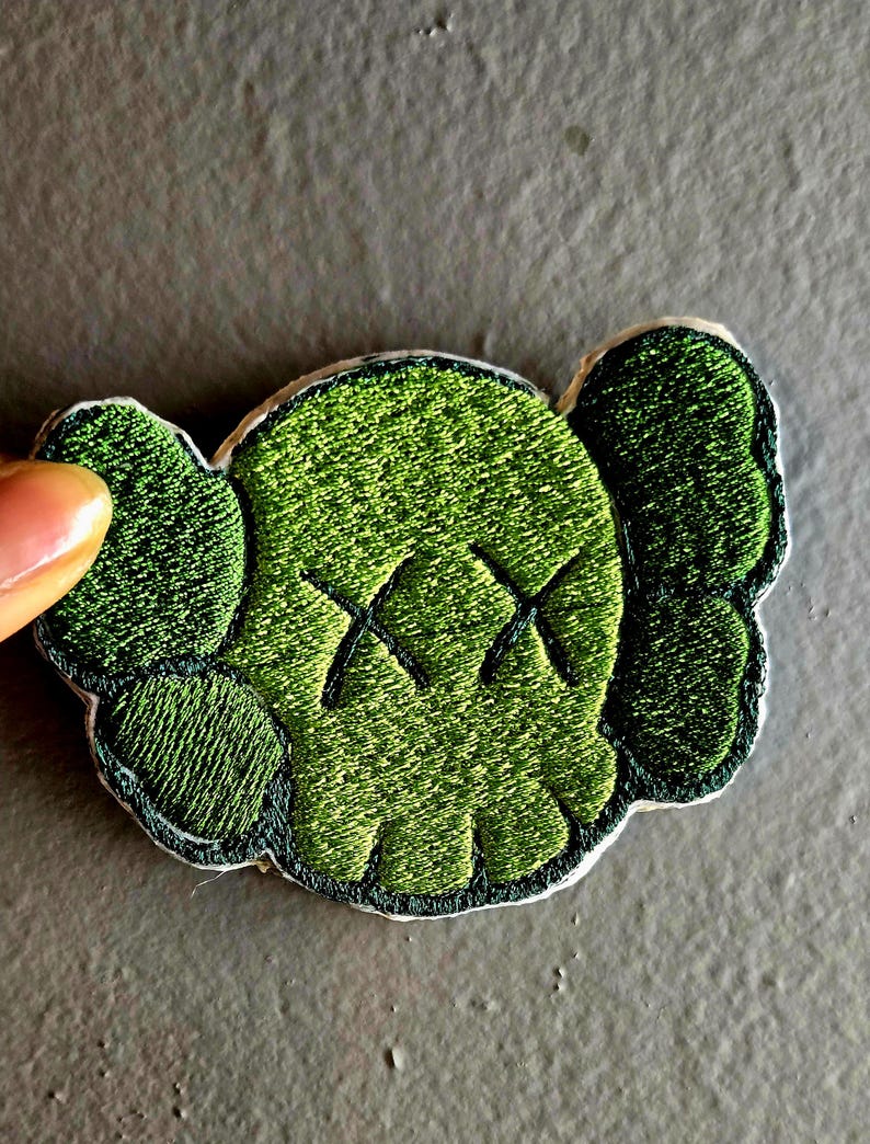 May include: Embroidered green patch featuring a stylized skull with X-shaped eyes and rounded ears. The patch has a dark green border and is set against a neutral background. The design is detailed with textured stitching.