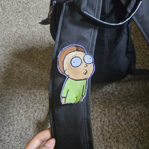 May include: A black backpack featuring an embroidered patch of a cartoon character with tan skin, brown hair, and a green shirt. The patch has a purple border. The backpack has a black handle and straps.