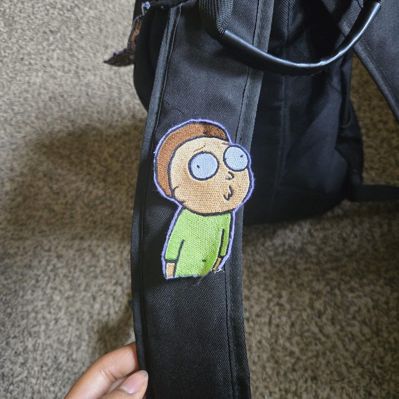 May include: A black backpack featuring an embroidered patch of a cartoon character with tan skin, brown hair, and a green shirt. The patch has a purple border. The backpack has a black handle and straps.