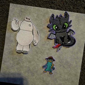 May include: Three embroidered patches featuring cartoon characters. One is a white and gray robot with a raised hand. Another is a black dragon with green eyes and purple accents. The third is a teal platypus wearing a brown hat.