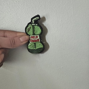 May include: Green embroidered patch with the words "WILD SNAKE" in red. The patch is in the shape of a stylized bottle or container with a black outline. The patch is held by a hand.
