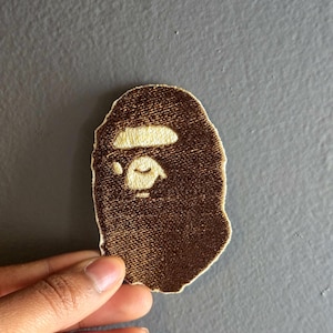 May include: Brown embroidered patch featuring a stylized ape face with a white eye and eyebrow. The patch has a textured appearance and is likely designed to be sewn or ironed onto fabric.
