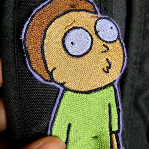 May include: Embroidered patch featuring a cartoon character with tan skin, brown hair, and large, light blue eyes. The character wears a green shirt and has a surprised expression. The patch is outlined in purple and is on a black fabric.