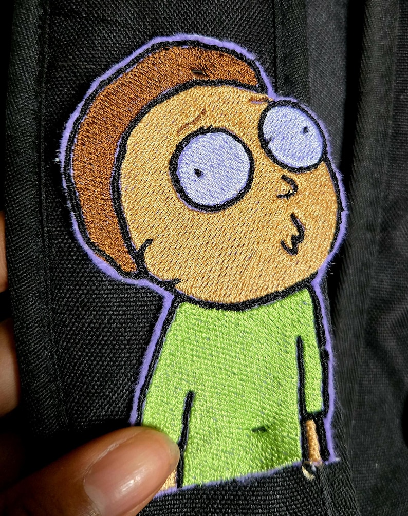 May include: Embroidered patch of a cartoon character with tan skin, brown hair, and large, light blue eyes. The character wears a green shirt and has a surprised expression. The patch is outlined in purple on black fabric.