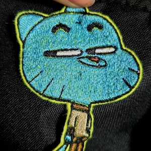 May include: Embroidered patch featuring a cartoon character with a blue head and body, yellow outline, and tan pants. The patch is attached to a black fabric. The character has black eyes and a small orange mouth.