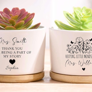 Può includere: Due vasi di ceramica bianca con piante grasse. Un vaso ha il testo "Mrs. Smith THANK YOU FOR BEING A PART OF MY STORY Sophia" e un cuore. L'altro vaso ha il testo "HELPING LITTLE MINDS GROW Mrs. Williams" e un cuore. Entrambi i vasi poggiano su sottovasi in legno.