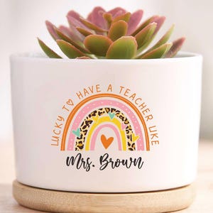 May include: White ceramic plant pot with a succulent. The pot features a rainbow design and the text "LUCKY TO HAVE A TEACHER LIKE" and "Mrs. Brown". It sits on a wooden base.