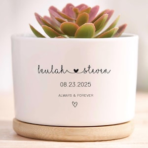 Può includere: Un vaso di ceramica bianco con una pianta succulenta. Il vaso ha una base in legno e presenta i nomi "Hulah & Steven", una data e le parole "Always & Forever" con un simbolo a cuore.