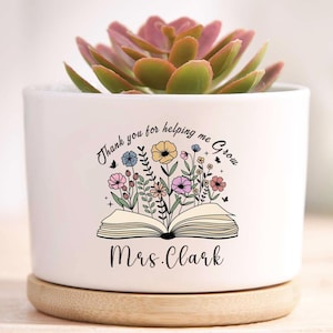 May include: White ceramic planter with a succulent plant. The planter has a wooden base and features the text "Thank you for helping me Grow" and "Mrs. Clark" with a floral book design. A thoughtful gift for a teacher.