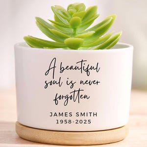 May include: A white ceramic planter with a green succulent plant. The planter has the text "A beautiful soul is never forgotten" and the name "JAMES SMITH 1958-2025". The planter sits on a wooden base.
