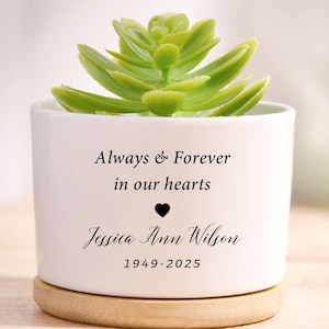Peut inclure: Un pot de fleurs en céramique blanche avec une plante succulente verte. Le pot porte l'inscription "Always & Forever in our hearts" avec un symbole de cœur et le nom "Jessica Ann Wilson 1949-2025". Le pot repose sur une base en bois.