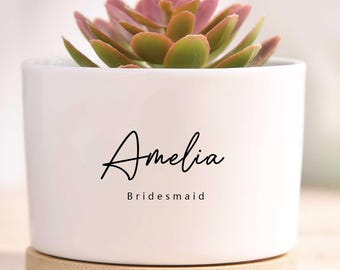 May include: White ceramic plant pot with a succulent. The pot has the name "Amelia" and the word "Bridesmaid" in black script. It sits on a round wooden base.