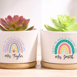 May include: Two white ceramic pots with succulents, each on a wooden saucer. The pots have a rainbow design and the text "LUCKY TO HAVE A TEACHER LIKE" with a teacher's name. One succulent is pink, the other is green.
