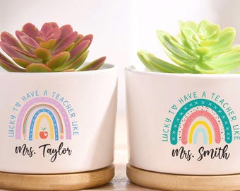 May include: Two white ceramic pots with succulents, each on a wooden saucer. The pots have a rainbow design and the text "LUCKY TO HAVE A TEACHER LIKE" with a teacher's name. One succulent is pink, the other is green.