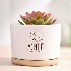 May include: A white ceramic planter with a succulent plant. The planter has the words "BESTIE est. 2020" and "AUNTIE est. 2026" in black script with red heart accents. The planter sits on a round, light-colored wooden base.