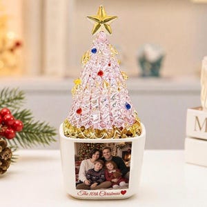 May include: A decorative tabletop Christmas tree ornament featuring a textured light pink crystalline cone topped with a gold star and accented with small colorful beads. The base is a white ceramic square planter displaying a family photo.