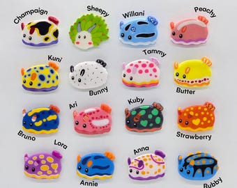 Puede incluir: Una colección de coloridos dijes decorativos con forma de babosas marinas. Cada dije presenta un diseño único con nombres como "Champaign", "Sheepy" y "Willani". Los dijes son de varios colores, incluyendo rosa, azul, amarillo y verde.