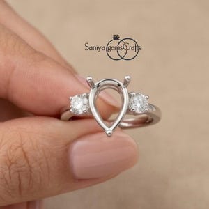 May include: A silver-toned metal ring setting featuring a central pear-shaped prong mount flanked by two round prong-set stones. The metal band is smooth and polished. The text Saniya gems Crafts is visible above the ring setting.