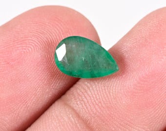 Puede incluir: Una piedra preciosa esmeralda tallada en forma de pera, de color verde intenso con inclusiones internas visibles, que presenta una superficie pulida que refleja la luz desde sus múltiples facetas angulares.