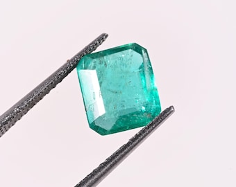 Puede incluir: Una esmeralda rectangular de tono verde vibrante con inclusiones internas visibles, sostenida por unas pinzas de metal oscuro sobre un fondo claro y liso. La piedra presenta un corte facetado con bordes definidos.