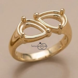 Könnte beinhalten: Ein goldener Ring mit zwei leeren birnenförmigen Fassungen in Bypass-Anordnung. Das Metallband ist glatt und poliert und für zwei tropfenförmige Edelsteine ausgelegt. Der Text Saniya gemsCrafts ist auf dem Ring zu sehen.