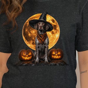 Op de afbeelding: Een donkergrijs gemêleerd T-shirt met korte mouwen en een grafische print van een Duitse staande hond met een zwarte heksenhoed en een oranje bandana, voor een grote volle maan met twee oplichtende pompoenen aan de onderkant.
