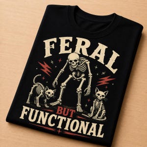 Könnte beinhalten: Schwarzes T-Shirt mit einem Skelett und zwei Katzen-Skeletten. Der Text lautet "FERAL" über dem Skelett und "BUT FUNCTIONAL" darunter. Rote Blitzgrafiken befinden sich an den Seiten des Skeletts.