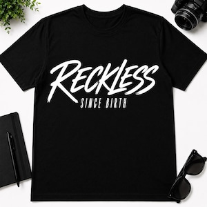 Op de afbeelding: Een zwart t-shirt met korte mouwen voorzien van de witte tekst RECKLESS in een groot, gestileerd penseelstreeklettertype, met de woorden SINCE BIRTH in een kleiner, strak schreefloos lettertype direct onder het hoofdontwerp op de borst.
