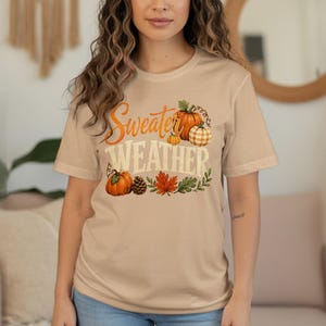 "Sweater Weather" graphic with pumpkins, pinecone, and autumn leaves on a tan t-shirt.
