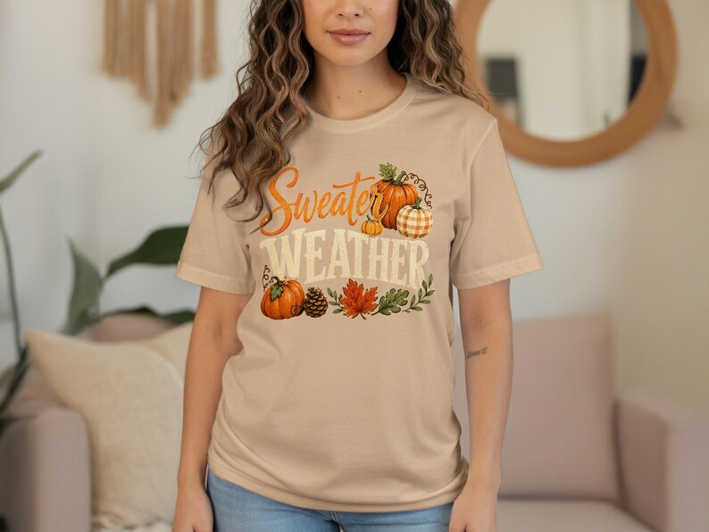 "Sweater Weather" graphic with pumpkins, pinecone, and autumn leaves on a tan t-shirt.