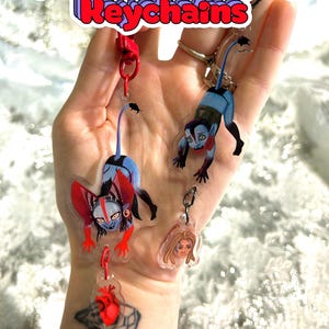 Puede incluir: Llaveros de acrílico transparente con figuras de dibujos animados y el texto "Quarang Hanging Keychains". Los llaveros presentan colores rojo, azul y negro. Un llavero tiene un diseño de corazón rojo. Los llaveros están sujetos a cierres rojos y plateados.