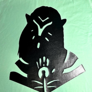 May include: A light green t-shirt featuring a large black graphic. The graphic is a stylized design of a creature with a hand and a crescent shape. The design is centered on the shirt.