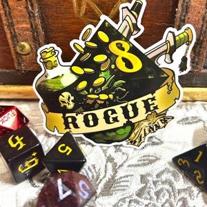May include: A sticker with a black and yellow design featuring a skull, coins, and crossed swords. The word "ROGUE" is written on a banner. Dice with yellow numbers are scattered around the sticker.