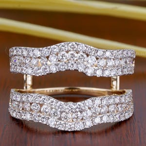 May include: A gold-coloured ring guard with a double-band design, set with many small, clear gemstones. The ring guard is displayed against a dark brown surface, with a blurred yellow and green background.