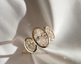 Può includere: Un anello d'oro con tre diamanti taglio ovale. I diamanti sono incastonati in una montatura a castone e l'anello è esposto su un tessuto bianco e setoso. Il testo "PRISHAJEWELSUUSA" è visibile.