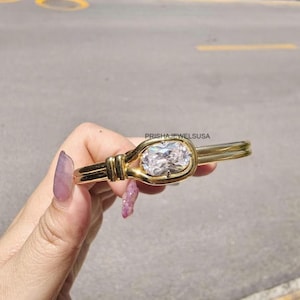 May include: A gold-toned bracelet featuring a large, clear, oval-shaped gemstone at its center. The bracelet has a unique knot-like design around the gemstone, with a smooth, polished finish. The brand name "PRISHAJEWELSUUSA" is visible.