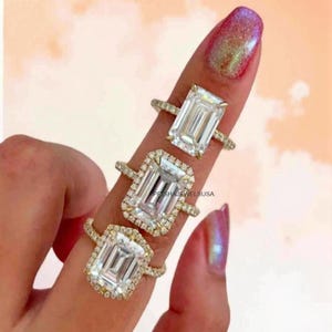 Peut inclure: Trois bagues en or avec de grandes pierres précieuses rectangulaires et transparentes. Deux bagues ont un halo de petites pierres autour de la pierre principale. Les bagues sont sur un doigt avec un vernis à ongles rose et irisé. Le texte "PRISHAJEWELSUUSA" est visible.