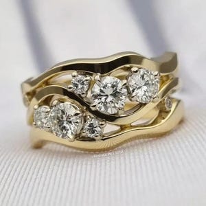 Puede incluir: Dos anillos de boda de oro con un diseño ondulado, cada uno engastado con múltiples diamantes redondos y claros. Los anillos están apilados, mostrando los intrincados detalles del engaste y el brillo de las piedras preciosas. El fondo es una tela blanca suave.