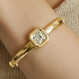 Peut inclure: Un bracelet jonc doré avec une pierre précieuse carrée et transparente au centre. La pierre est sertie dans une lunette dorée de forme octogonale. Le bracelet est lisse et poli, avec le texte "PRISHAJEWELSUSA" visible.
